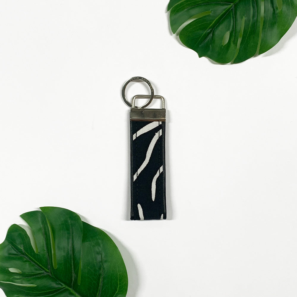 a photo of a keyfob made of batik in the pattern black fern against a neutral background surrounded by two tropical leaves