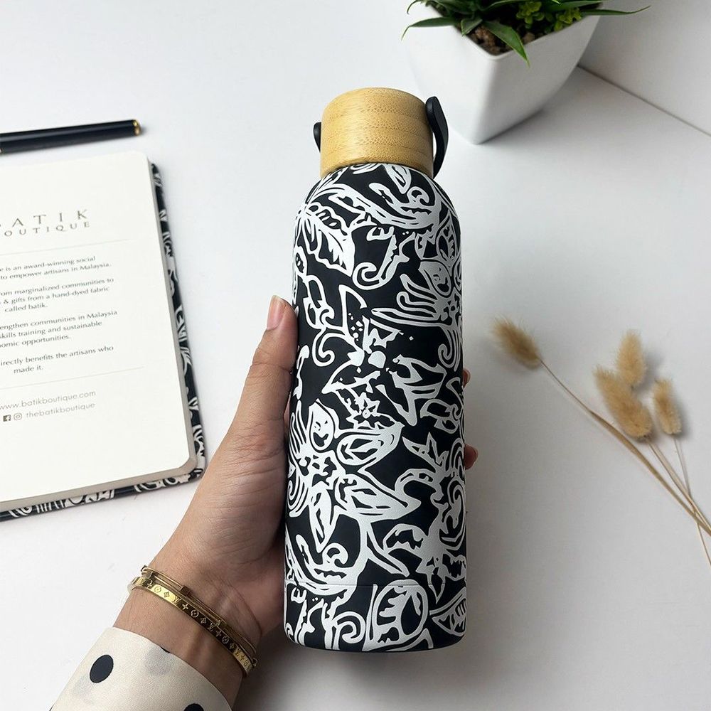 Hand holding a black and white patterned water bottle with a wooden cap on a white surface.