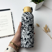 Hand holding a black and white patterned water bottle with a wooden cap on a white surface.