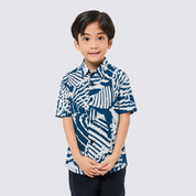 A young boy wearing a short-sleeve button-up shirt with a blue and white abstract batik pattern, standing against a light gray background.