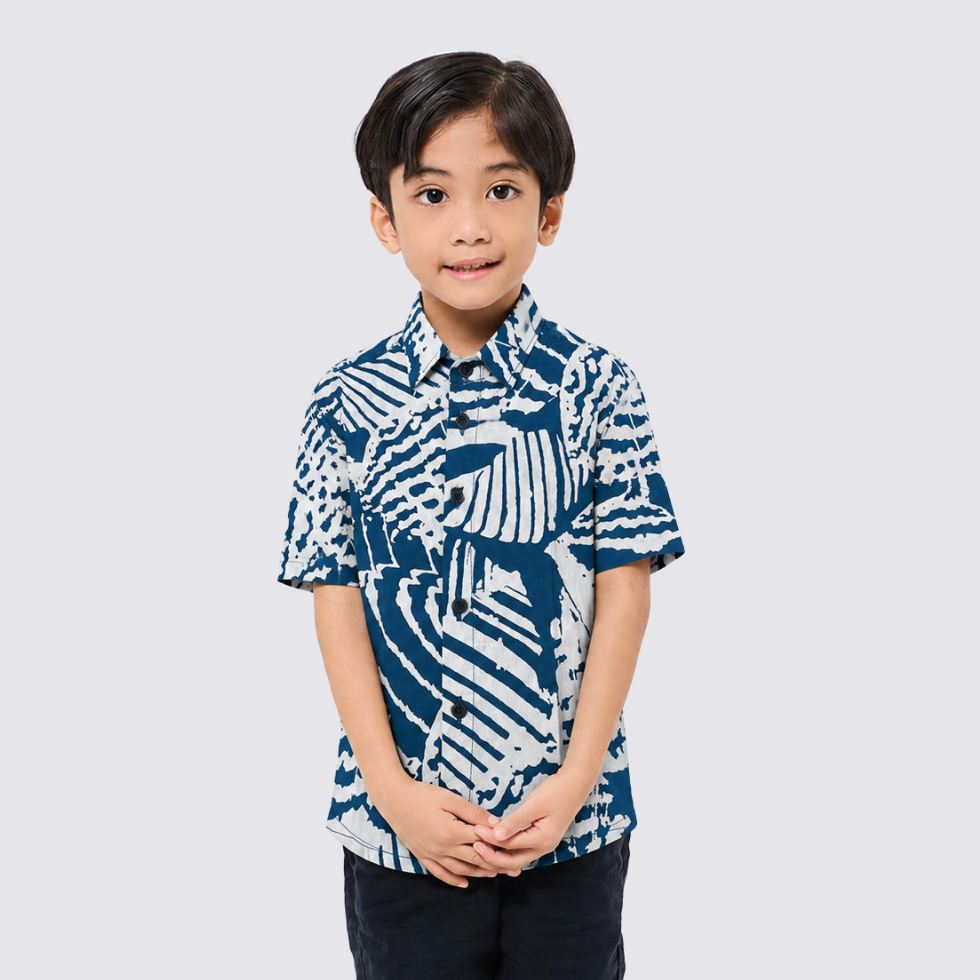 A young boy wearing a short-sleeve button-up shirt with a blue and white abstract batik pattern, standing against a light gray background.