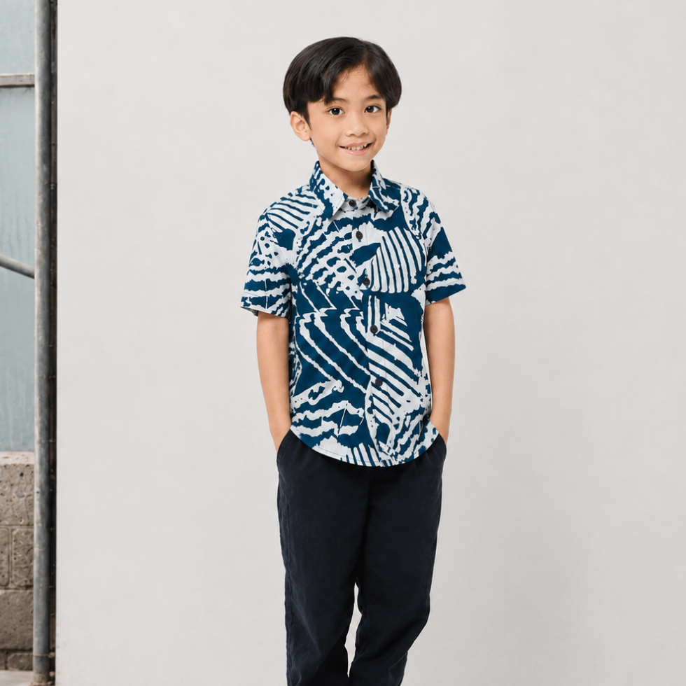 A young boy wearing a short-sleeve button-up shirt with a navy blue and white batik-style abstract pattern, paired with dark pants, standing against a plain light gray wall.