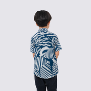 A child viewed from behind wearing a short-sleeve button-up shirt with a bold blue and white abstract graphic print, paired with dark pants, against a light gray background.
