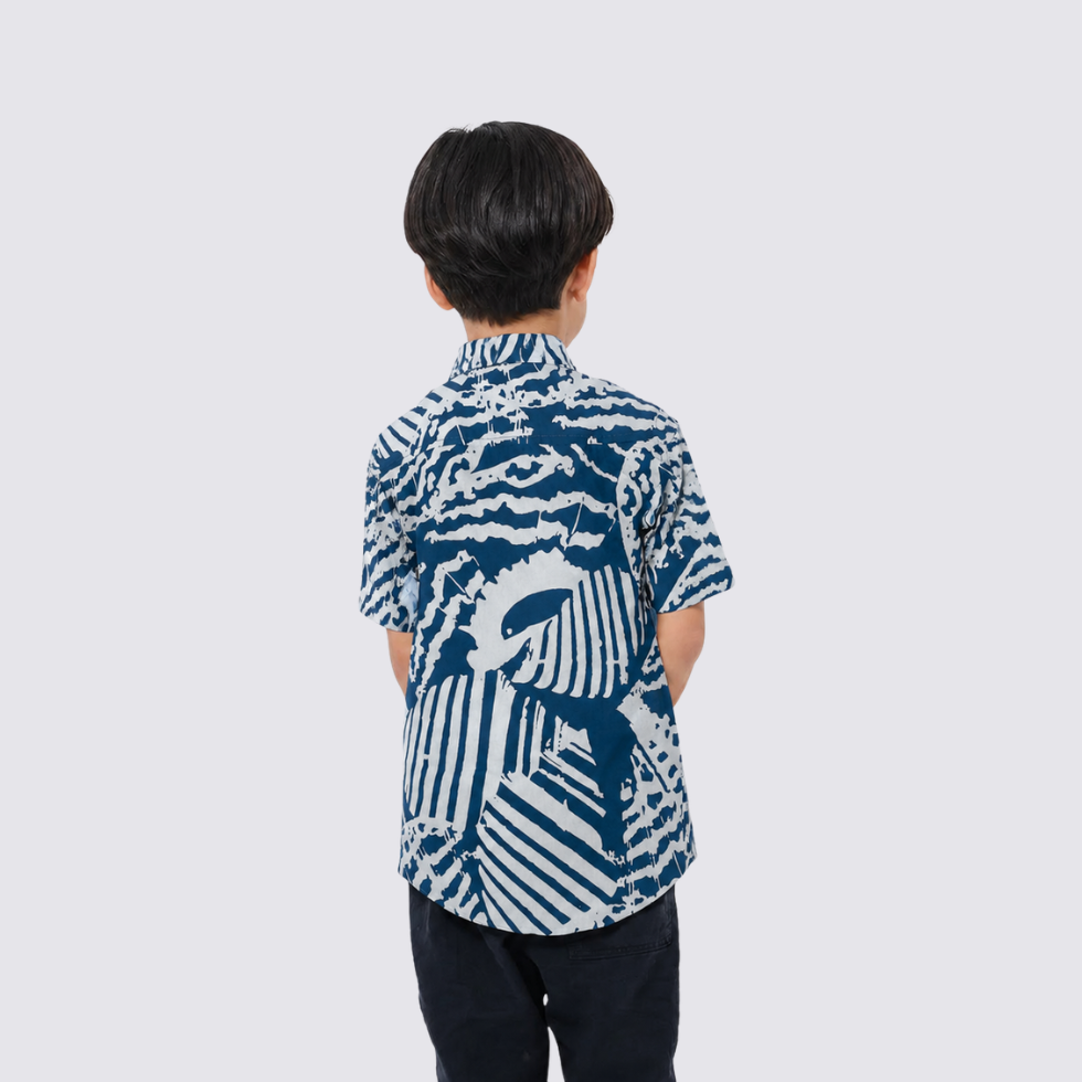 A child viewed from behind wearing a short-sleeve button-up shirt with a bold blue and white abstract graphic print, paired with dark pants, against a light gray background.