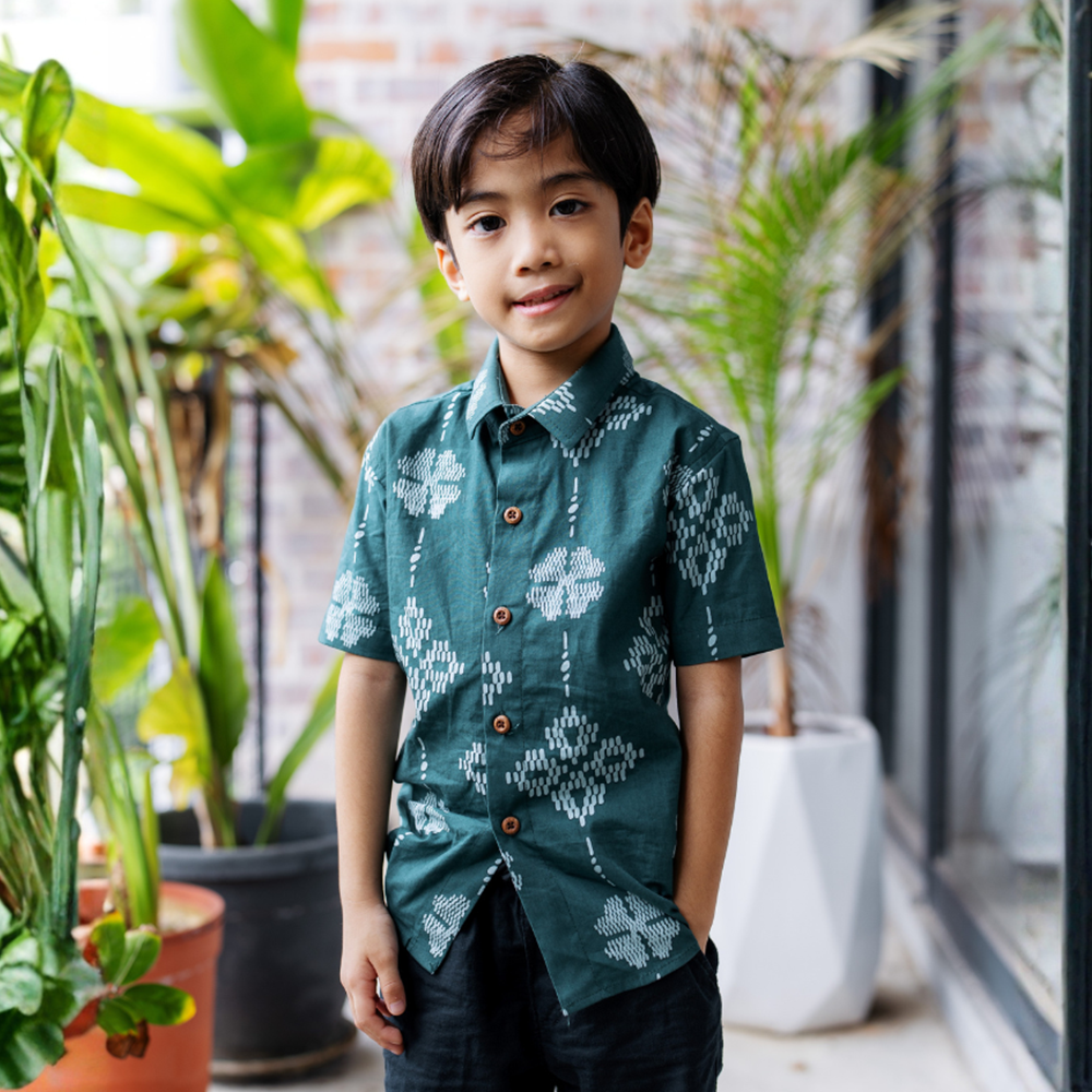 Child wearing a green patterned shirt standing among potted plants