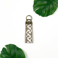 a picture of brown arabesque key fob made of batik remnants against a neutral background with tropical leaves as decoration