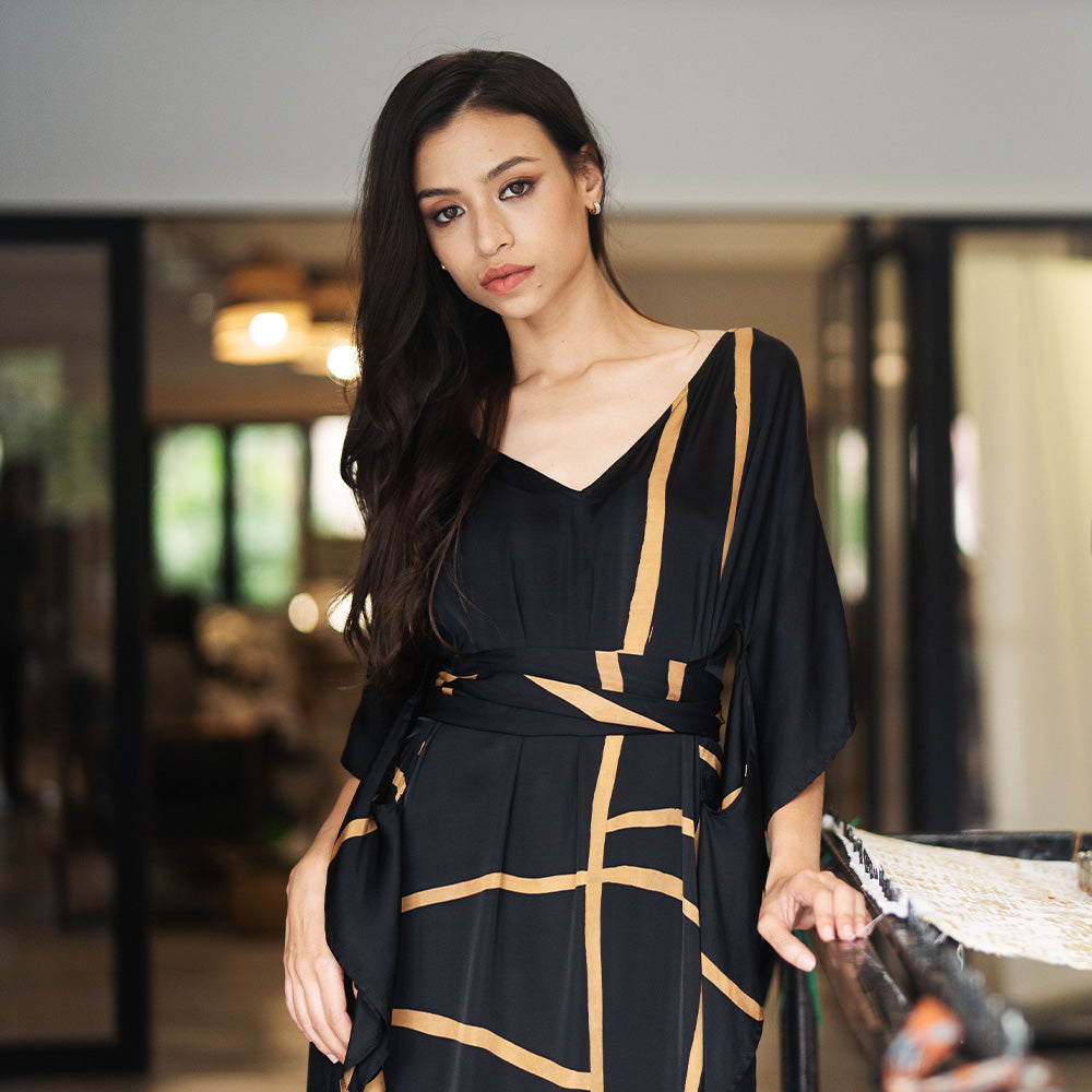 a lifestyle photo of model wearing batik caftan dress in black ecru pattern standing next to frame for batik making