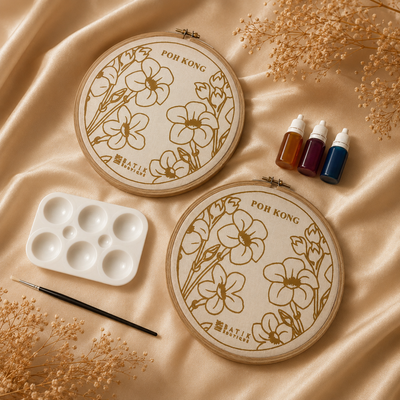 Two embroidery hoops with floral patterns, paint bottles, an palette, and a brush on a beige fabric background.
