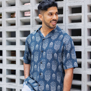 a lifestyle photo of man leaning by wall of brick wearing batik cuban shirt in navy pineapple pattern