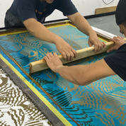 Two people pressing a large blue and gold silk screen with a wooden roller in a workshop setting.