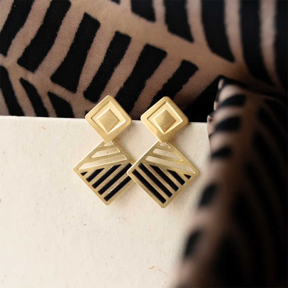 Close-up of gold geometric earrings with cut-out detailing, placed on a textured surface, paired with batik fabric in the background.