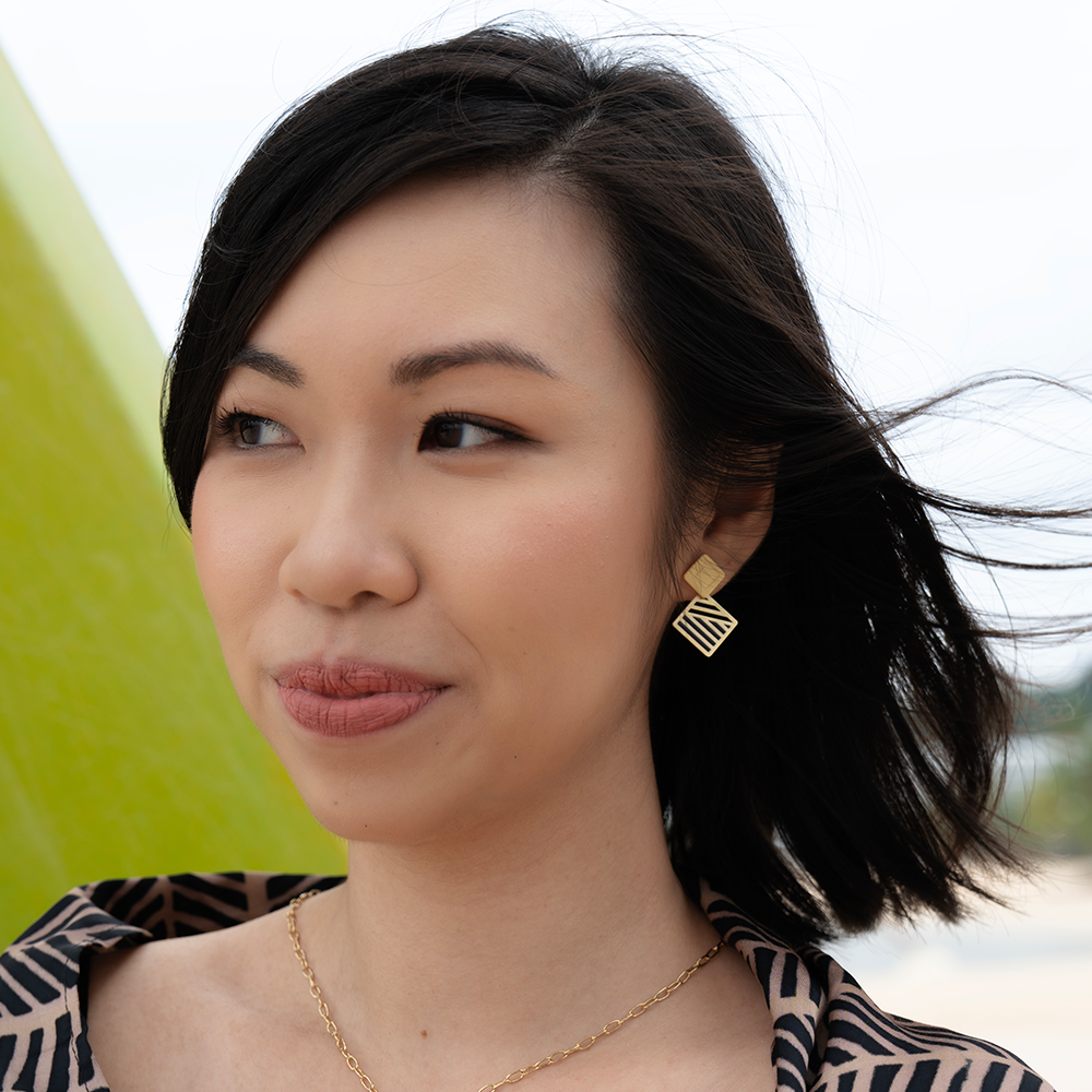 Woman wearing gold geometric earrings with short black hair, styled in a batik top, outdoors with a natural background.