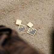 Gold geometric earrings with black enamel details resting on sandy beach surface, partially framed by batik fabric.