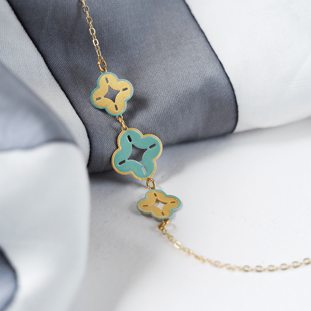 Gold bracelet with three batik-inspired floral charms in turquoise and gold, laid on folded grey and white fabric.