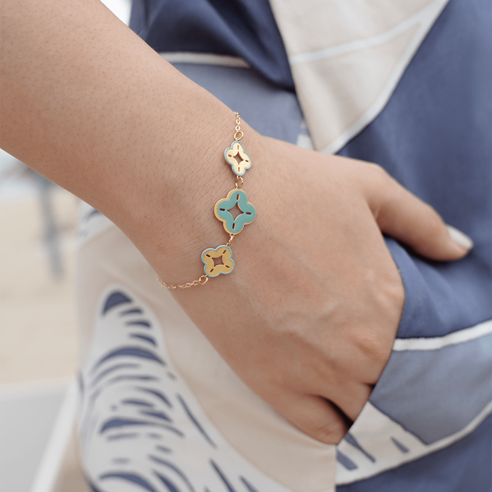 Close-up of a woman's wrist wearing a turquoise and gold floral charm bracelet with a beach background.