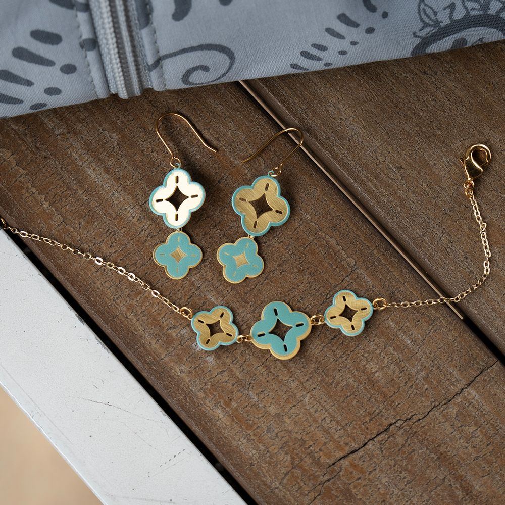 Lifestyle shot of gold and turquoise batik bracelet and earrings set on a wooden surface with grey batik fabric.