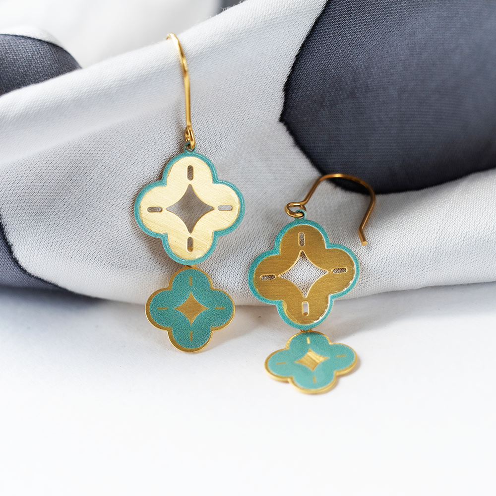 Gold geometric square earrings with cut-out patterns, placed on a textured background with batik cloth.