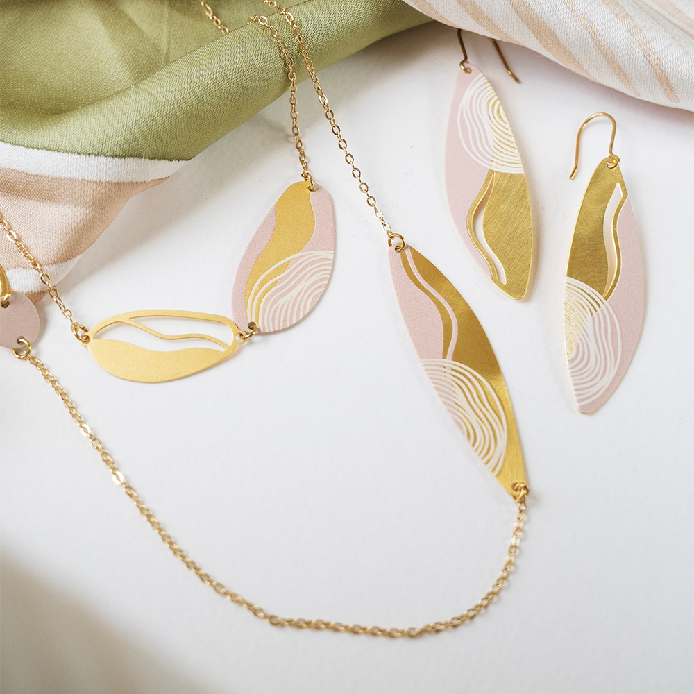 Flat lay of matching batik-inspired earrings and necklace in matte gold and pink on a fabric backdrop.