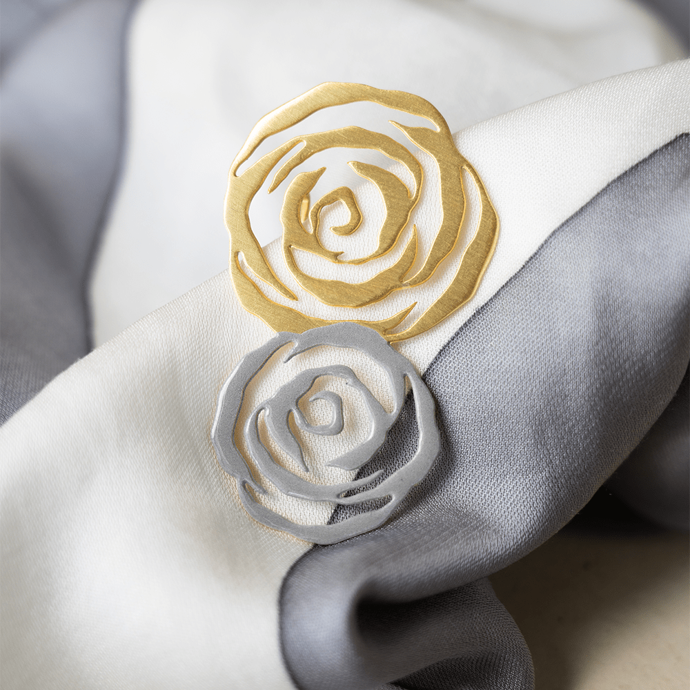 Close-up of two abstract rose-shaped brooches in matte gold and silver finishes, styled on a soft, folded white and grey fabric background.
