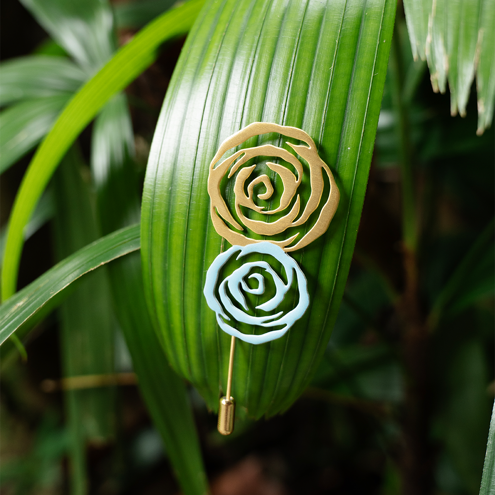 Gold and silver rose brooches pinned to a green tropical palm leaf, showcasing their contrast and intricate rose petal design.