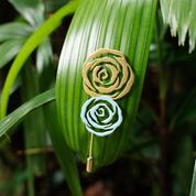 Gold and silver rose brooches pinned to a green tropical palm leaf, showcasing their contrast and intricate rose petal design.