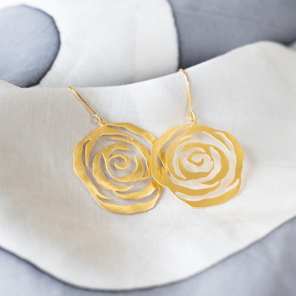 A pair of gold rose-shaped earrings resting on soft white and grey batik fabric.