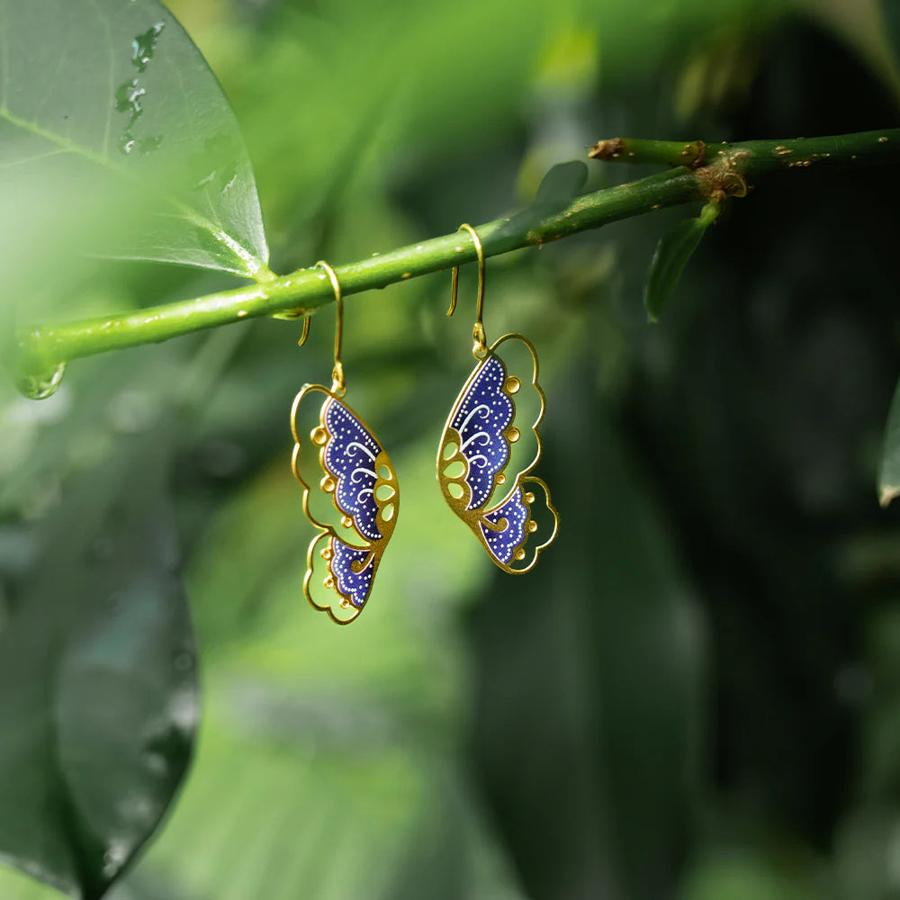 Butterfly-shaped earrings with blue and gold design hanging on a green leaf.