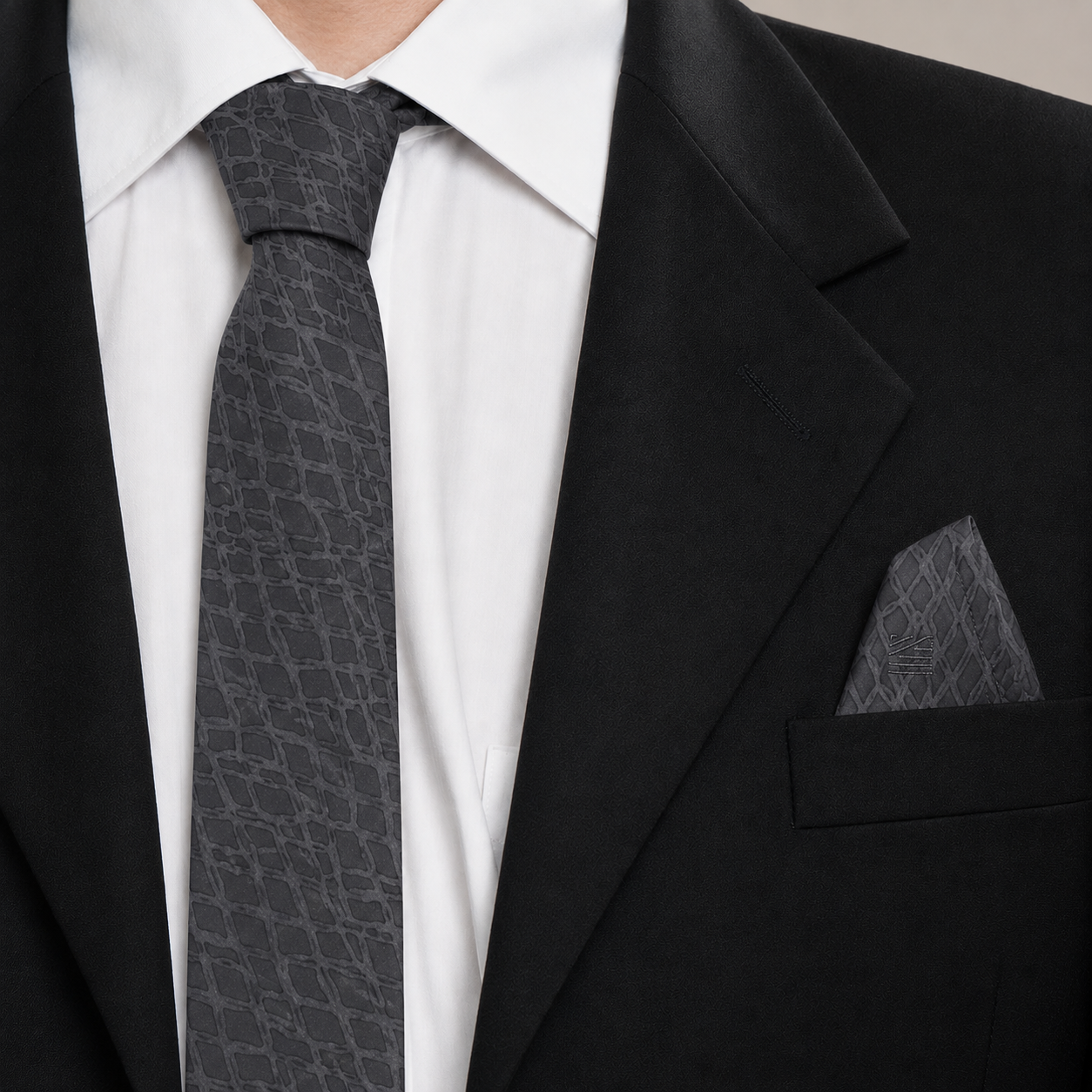 A close-up of a men's formal outfit featuring a dark gray patterned silk tie and matching pocket square, styled with a white dress shirt and a black suit jacket.
