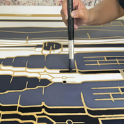 A hand holding a black paintbrush applies dark navy and white patterned material onto a large sheet with gold linework, likely wallpaper or a decorative wall covering.