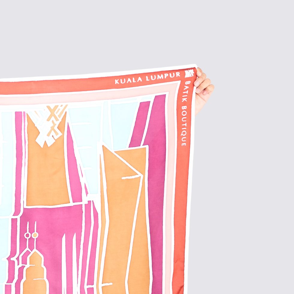 A hand holds up a square scarf featuring a colorful abstract cityscape design with the text 'KUALA LUMPUR BIK BOUTIQUE' and 'BAIK BOUTIQUE' printed along the border, set against a solid light gray background.