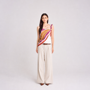 A woman stands against a light gray studio background wearing a sleeveless wrap-style top with diagonal striped panels in white, orange, magenta, and burgundy, paired with wide-leg beige trousers and brown pointed shoes.