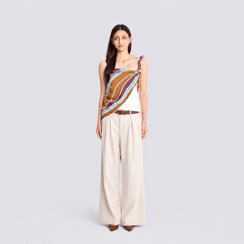 A woman stands against a light gray studio background wearing a sleeveless wrap-style top with diagonal striped panels in white, orange, magenta, and burgundy, paired with wide-leg beige trousers and brown pointed shoes.