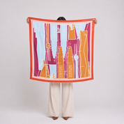 A person stands against a light gray studio background holding up a square scarf. The scarf features a stylized cityscape illustration with orange, magenta, light blue, and white elements, framed by a bright orange border.