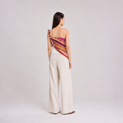 A woman stands with her back to the camera wearing a one-shoulder wrap top with a colorful striped pattern in red, orange, yellow, and purple, paired with high-waisted cream wide-leg trousers.