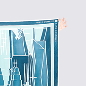 A hand holds the top-right corner of a printed map-style scarf or poster featuring blue and white city building illustrations. The printed text reads 'KUALA LUMPUR' and 'BANK BOUTIQUE'.