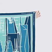 A hand holds the top-right corner of a printed map-style scarf or poster featuring blue and white city building illustrations. The printed text reads 'KUALA LUMPUR' and 'BANK BOUTIQUE'.