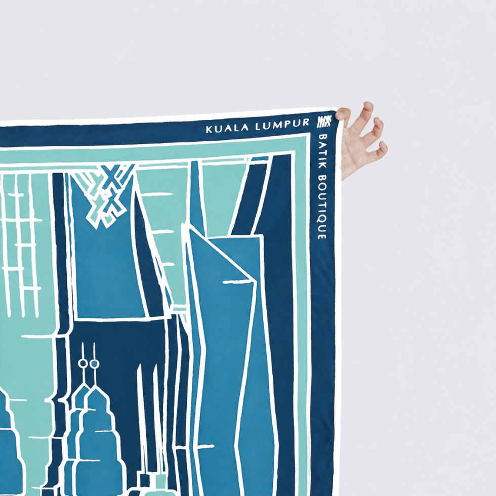 A hand holds the top-right corner of a printed map-style scarf or poster featuring blue and white city building illustrations. The printed text reads 'KUALA LUMPUR' and 'BANK BOUTIQUE'.