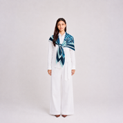 A woman stands against a light gray studio background wearing a white long-sleeve top and wide-leg white pants, with a patterned scarf draped around her shoulders in shades of blue, teal, and navy.