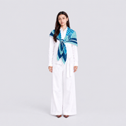 A woman stands against a light gray studio background wearing a white long-sleeve top and wide-leg white pants, with a patterned scarf draped around her shoulders in shades of blue, teal, and navy.