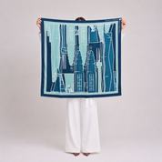 A person stands facing away from the camera holding up a square scarf. The scarf features a graphic illustration of Parisian-style buildings in shades of navy, teal, and light blue with white outlines.