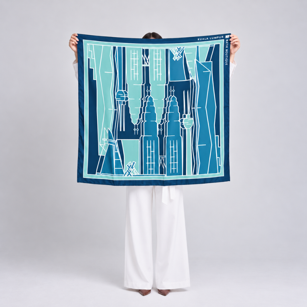 A person stands facing away from the camera holding up a square scarf. The scarf features a graphic illustration of Parisian-style buildings in shades of navy, teal, and light blue with white outlines.