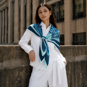 A woman wearing a white long-sleeve shirt and white pants, with a blue and teal patterned silk scarf tied around her neck, leaning against a stone wall in front of a brick building.