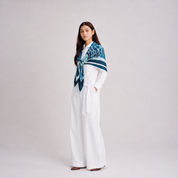 A woman stands in a studio wearing a white long-sleeve top and wide-leg white pants, with a blue and teal patterned scarf wrapped around her shoulders.