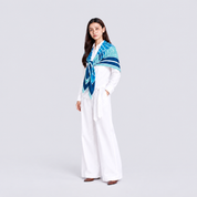 A woman stands in a studio wearing a white long-sleeve top and wide-leg white pants, with a blue and teal patterned scarf wrapped around her shoulders.