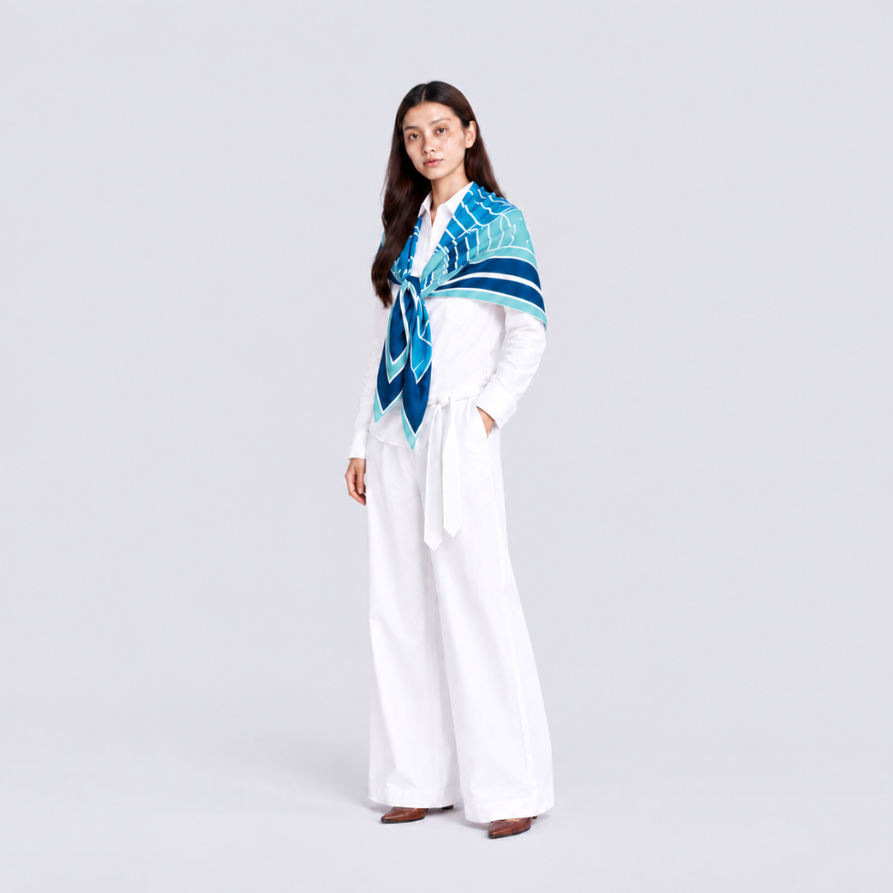 A woman stands in a studio wearing a white long-sleeve top and wide-leg white pants, with a blue and teal patterned scarf wrapped around her shoulders.