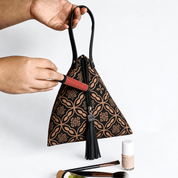 A black and brown patterned handbag with a tassel is held by two hands, with a lipstick being applied to the zipper. Makeup brushes and a nail polish bottle are visible on a white background.