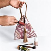 A maroon and gold patterned handbag with a tassel zipper is held by two hands, with cosmetic items placed near the bottom of the frame on a light gray background.