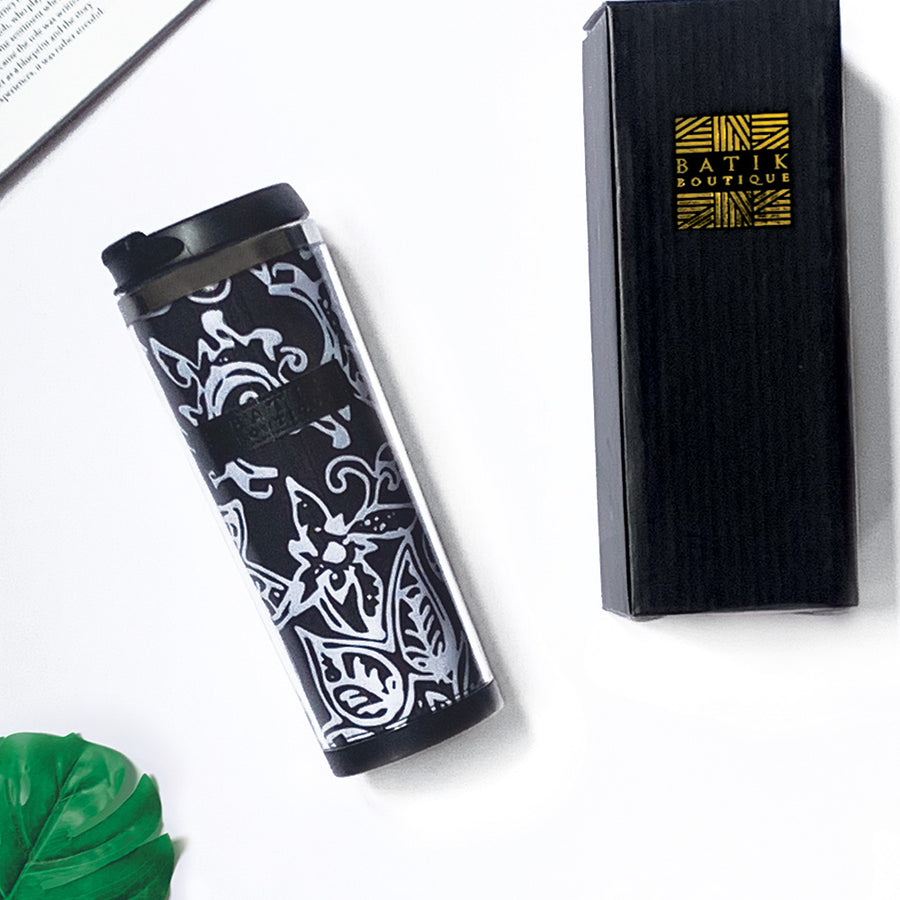 a lifestyle photo of a batik tumbler in the pattern black bunga against a neutral background and a black box, tropical leaf and a book as decorations