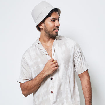 man posing in front of a white wall while wearing a shibori cuban shirt in mangosteen