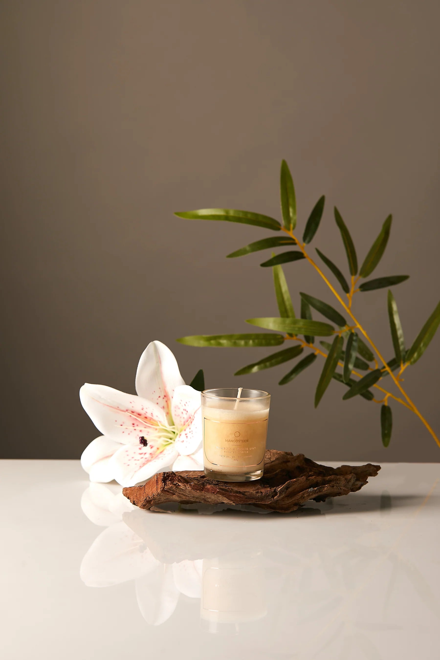 Candle in a glass holder with a white flower and green leaves on a reflective surface.